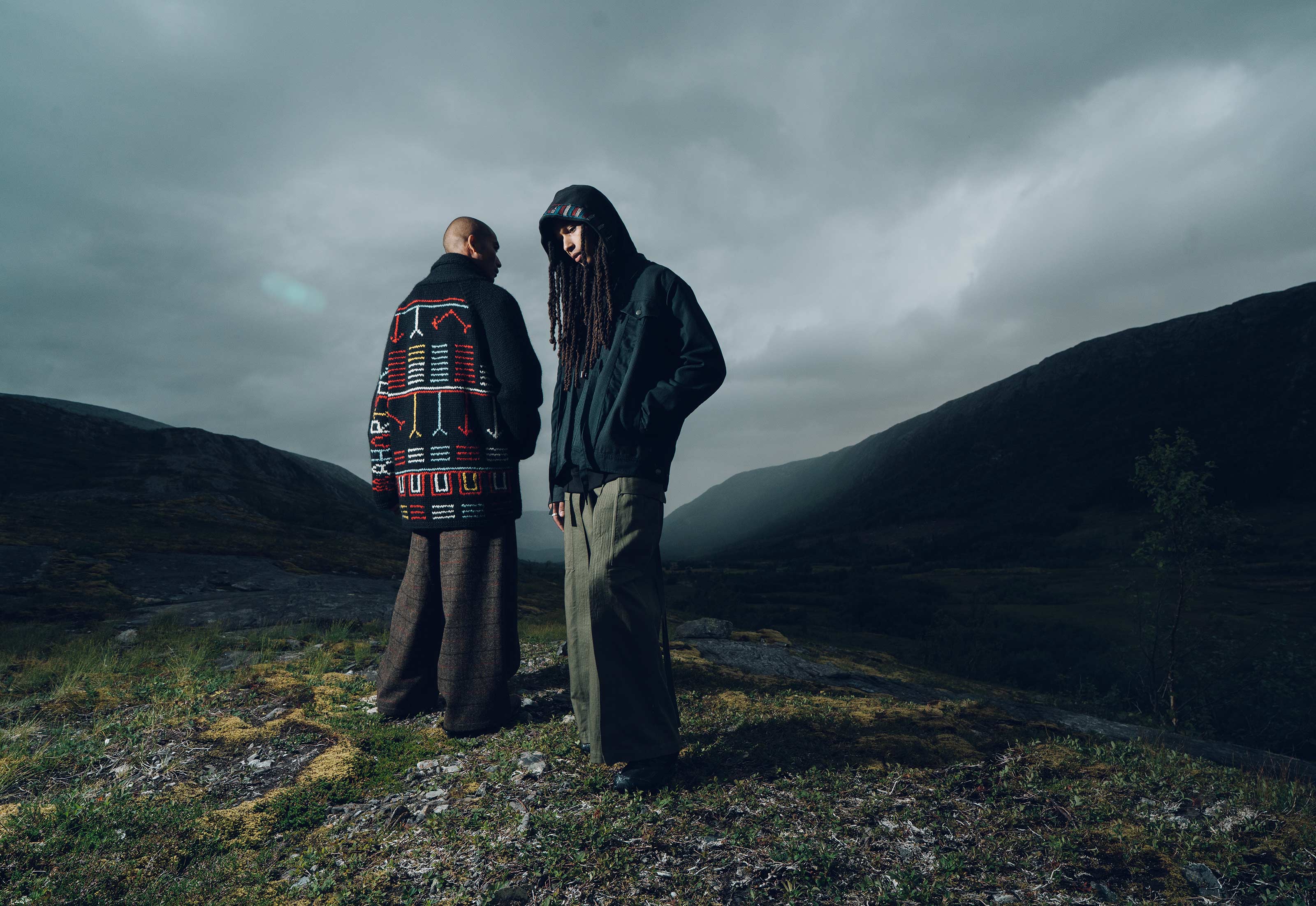 Two people standing in a mountainous landscape with a dark, moody sky.
