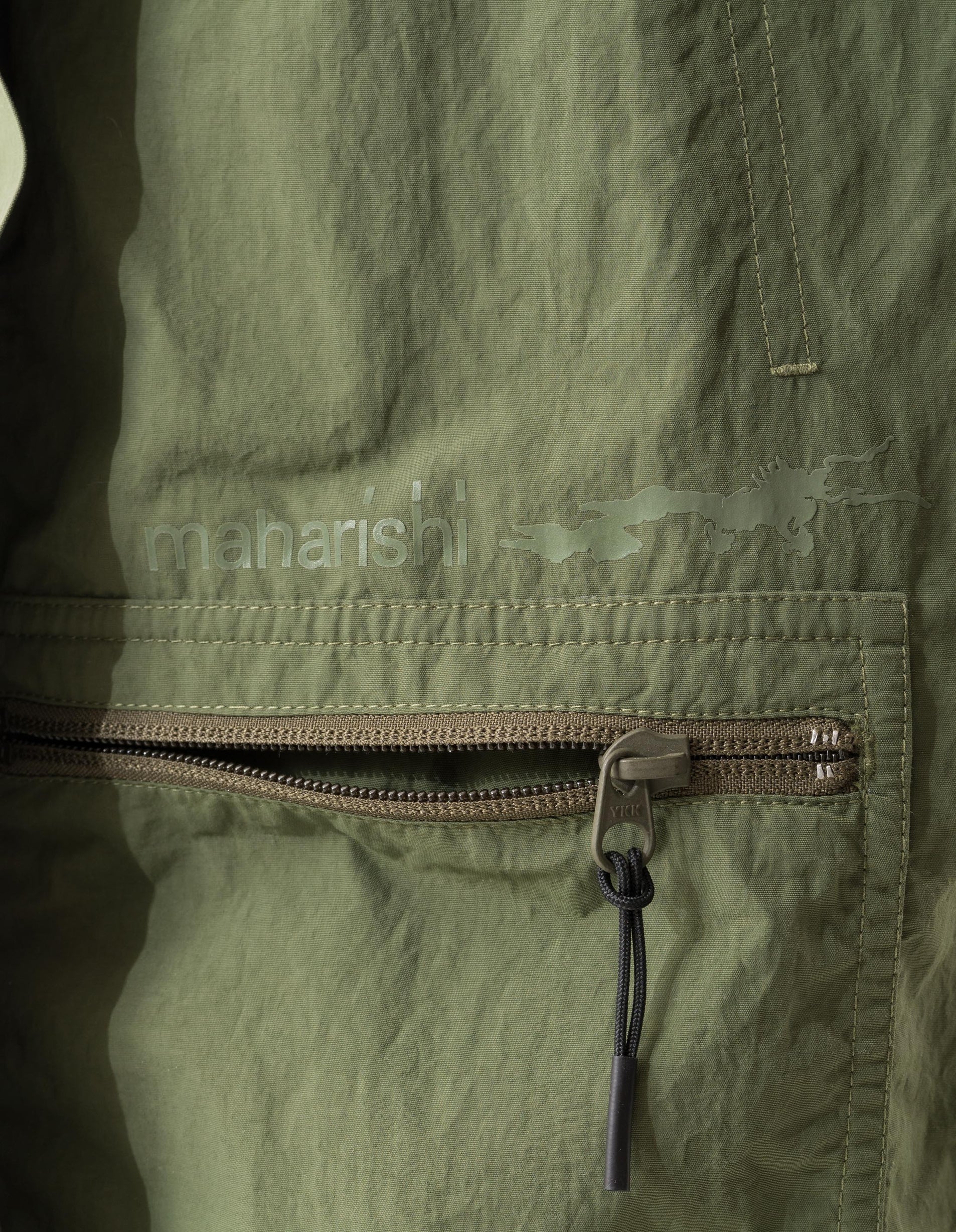 Close-up of a green jacket with a zipper, featuring the brand 'maharishi'.