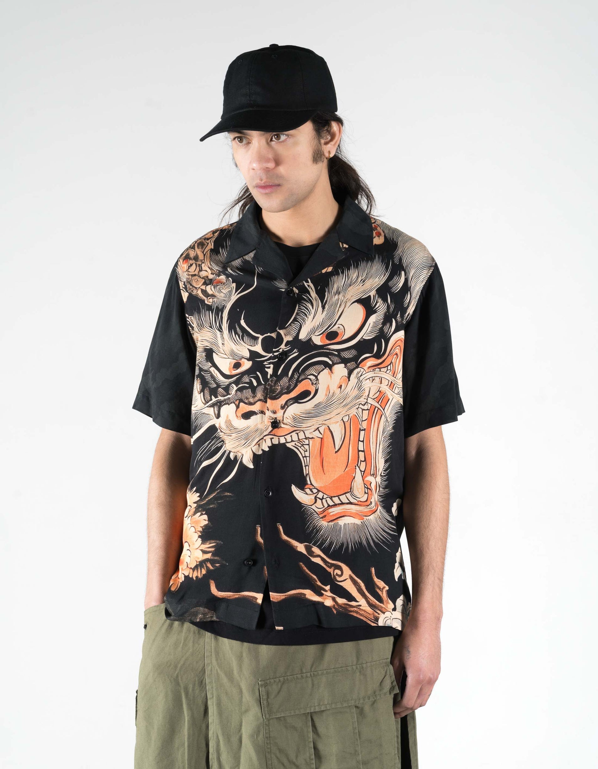 Person wearing a black shirt with a dragon design and a black cap on a white background